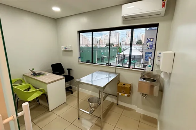 Consultório com mesa de atendimento, cadeiras, mesa de exame de inox e pia, iluminado por janela ampla