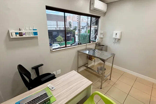 Consultório com mesa de exame de inox, mesa de trabalho com notebook e prateleiras de apoio