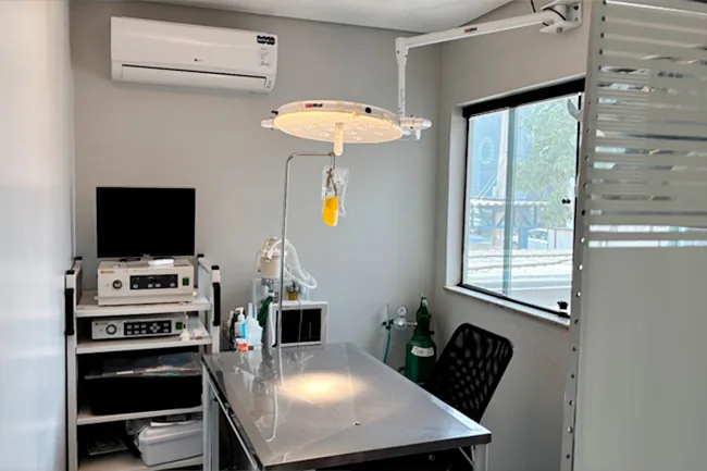 Sala de cirurgia com mesa de inox, foco cirúrgico de teto, aparelho de anestesia inalatória e janela lateral