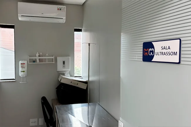 Sala de ultrassom, com placa de identificação na parede e mesa de exame de inox