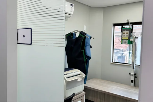 Sala de RX identificada na parede, com aventais de chumbo e equipamento de radiografia
